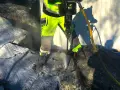 Arbetare med reflexkläder borrar i berg med en kraftig borrhammare. Dammet yr runt borren.
