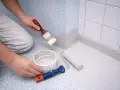 Person målar våtrumsmatta med pensel och roller. En hink med färg står bredvid.