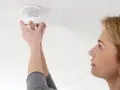 En kvinna installerar en brandvarnare. Dessa brandvarnare som pratar med varandra är viktiga för säkerheten.
