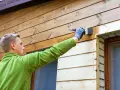 Man målar tryckimpregnerat trä med en pensel. Han har en grön tröja och grå handskar.