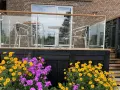 Vacker altan med glasräcke och trädetaljer. Blommor i gult och lila ramar in staketet. Perfekt altan staket inspiration för en modern uteplats.