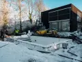 Snö täcker byggarbetsplatsen där en utbyggnad av husets långsida pågår. En grävmaskin och en dumper syns vid en nybyggd poolgrund.