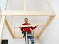 Man på stege bygger ett loft av trä. Han håller en träbit och ler.