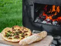 En pizza och ett bröd ligger på en skärbräda bredvid en pizzaugn med flammande ved.