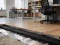Detaljerad vy av ljudisolering golv med trägolv ovanpå. Ett kontor med möbler syns i bakgrunden.