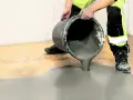 Person i varselkläder häller flytspackel golv ur en hink. Det gråa spacklet sprider sig över det gamla golvet.