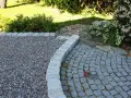 En vacker grus uppfart kantad av sten leder till en stenlagd uteplats. Grönska och buskar ramar in området.