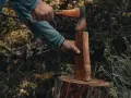 En person förbereder sig för att kapa ved utomhus. En yxa är redo att slå mot en träbit som står på en stubbe.