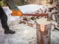 Person med yxa klyva ved i snöig skog. En hög med ved i bakgrunden.