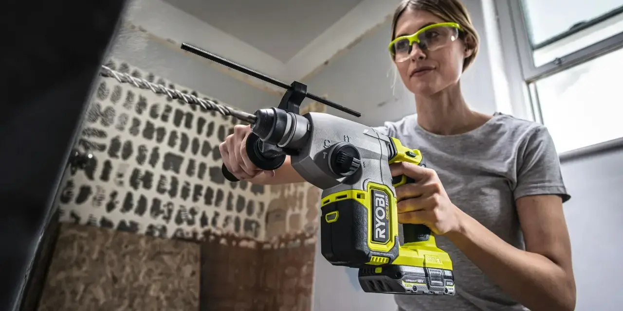 Kvinna med skyddsglasögon borrar i betongtak med en kraftfull slagborrmaskin. Damm virvlar upp från arbetet.