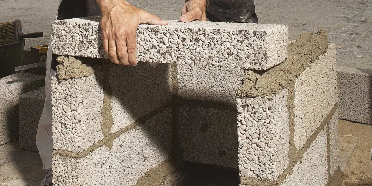 Händer lägger ett mura lecablock på en grund av cement. Byggnationen pågår utomhus.