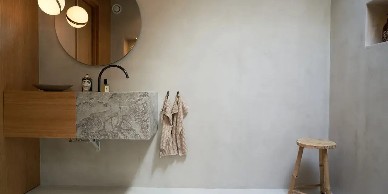 Modernt badrum med handfat i marmor och väggar av microcement. Enkel träpall och rund spegel.