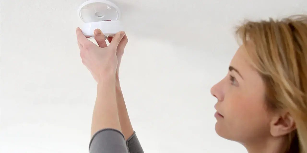En kvinna installerar en brandvarnare. Dessa brandvarnare som pratar med varandra är viktiga för säkerheten.