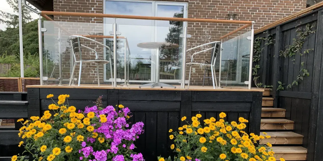 Vacker altan med glasräcke och trädetaljer. Blommor i gult och lila ramar in staketet. Perfekt altan staket inspiration för en modern uteplats.