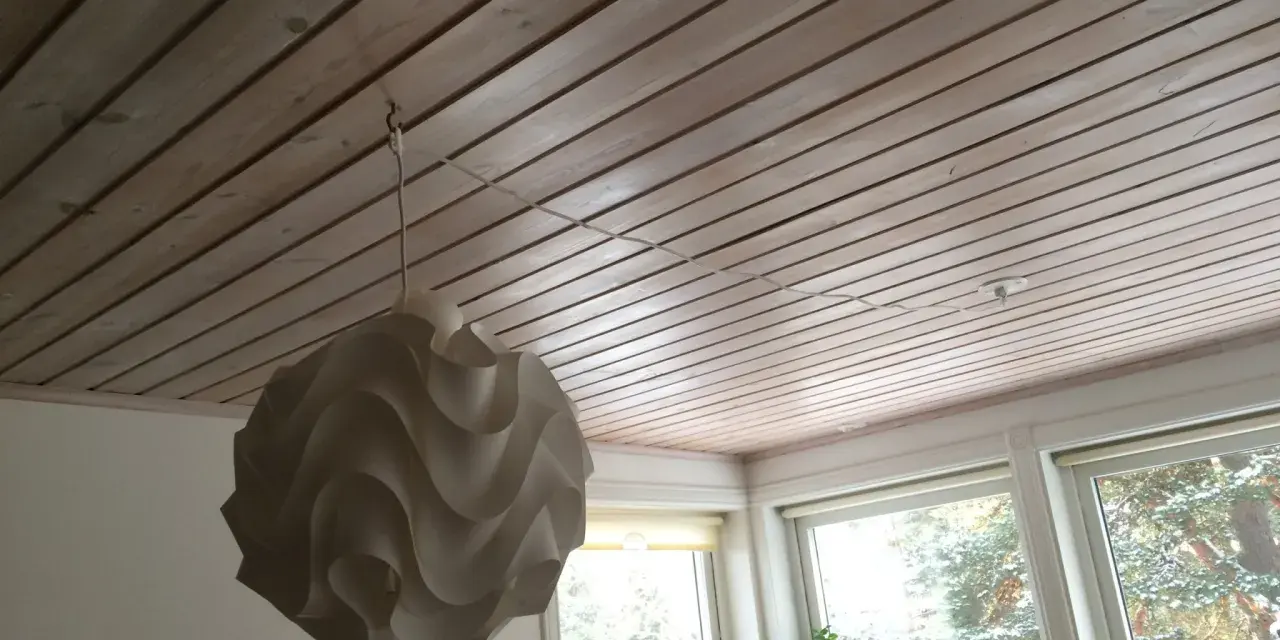 En vit taklampa hänger från en krok i ett träpanelat tak. En tunn sladd löper från lampan längs taket mot ett uttag.