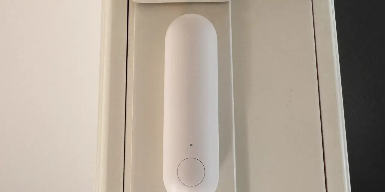 En vit, avlång sensor med en knapp sitter monterad på en ljusbeige dörrkarm. Den är en del av ett Aqara Thread-system för smart hem.