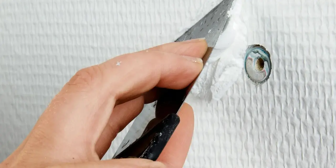 Hand spacklar ett litet hål i en vit, strukturerad vägg med en spackelspade.