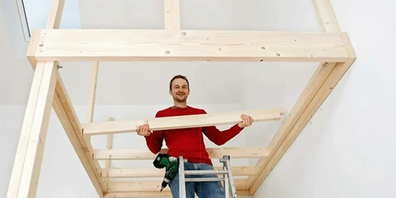 Man på stege bygger ett loft av trä. Han håller en träbit och ler.