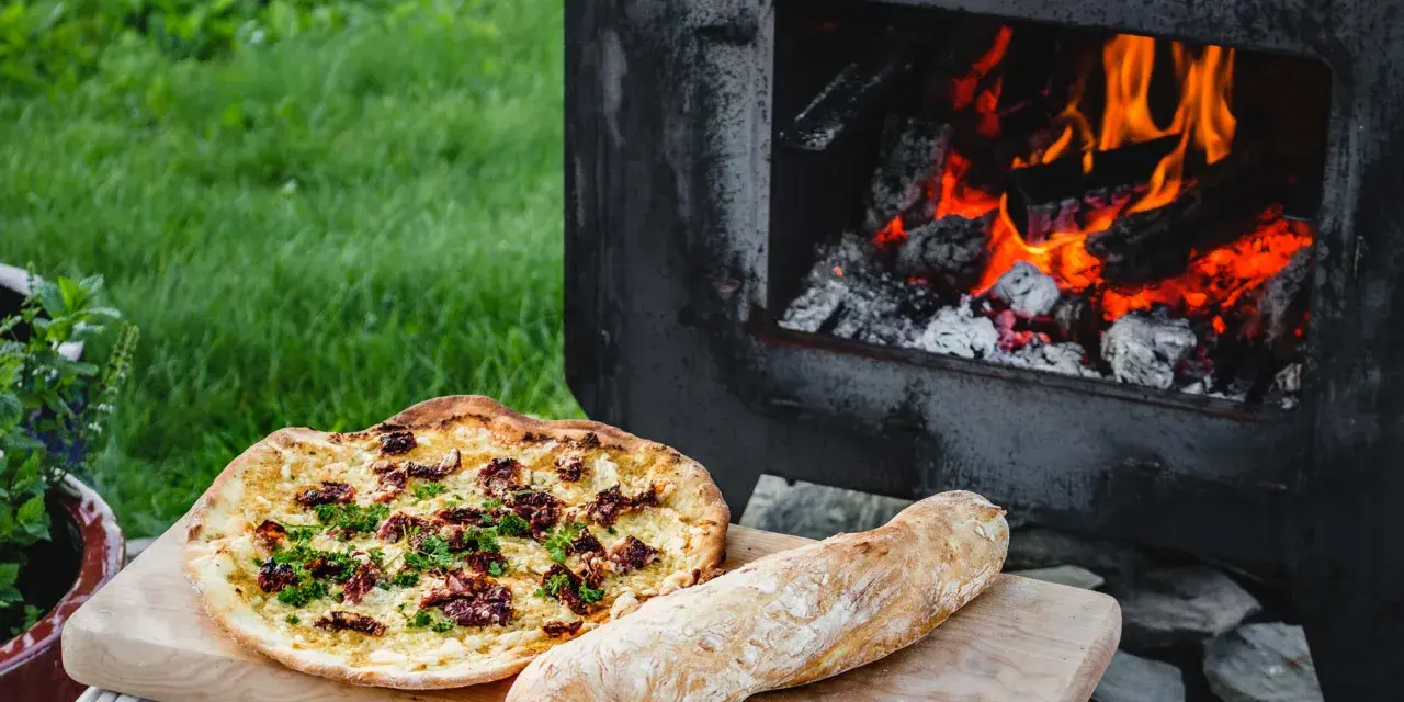 En pizza och ett bröd ligger på en skärbräda bredvid en pizzaugn med flammande ved.