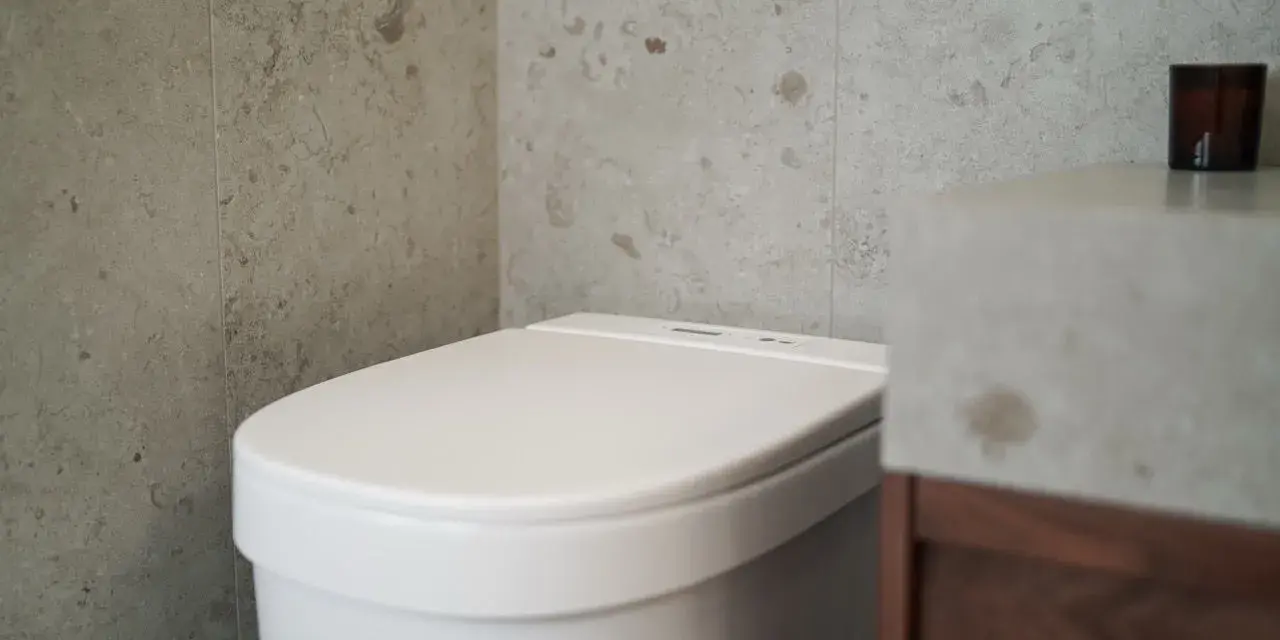 Modernt badrum med en vit toalett utan avlopp, grå kaklade väggar och ett handfat i betong.