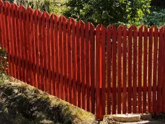 Rött trästaket med spetsiga toppar ger fin staket inspiration. Grönskande träd i bakgrunden.