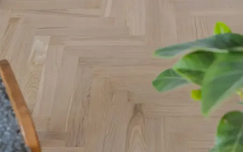 Ett nyslipat parkettgolv i fiskbensmönster, redo att skyddas. En grön växt och en del av en stol syns i bild.