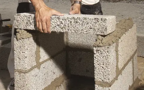 Händer lägger ett mura lecablock på en grund av cement. Byggnationen pågår utomhus.