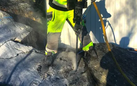 Arbetare med reflexkläder borrar i berg med en kraftig borrhammare. Dammet yr runt borren.