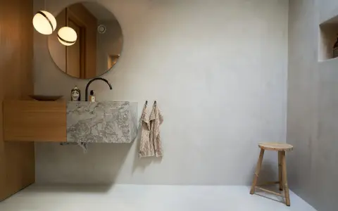 Modernt badrum med handfat i marmor och väggar av microcement. Enkel träpall och rund spegel.