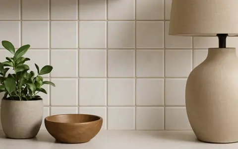 En krukväxt, en träskål och en lampa står framför en vägg med ljusa, fyrkantiga kakelplattor. En fin detalj som visar skillnad på kakel och klinker.