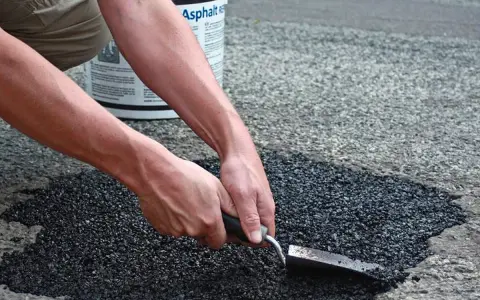 Händer som reparerar asfalt med en spade. En hink med "Asphalt" syns i bakgrunden.