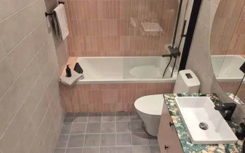 Ett modernt kaklat badrum med rosa kakel vid badkaret och grått klinkergolv. En vit toalett och ett handfat med en unik bänkskiva.