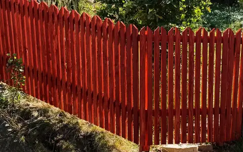 Rött trästaket med spetsiga toppar ger fin staket inspiration. Grönskande träd i bakgrunden.