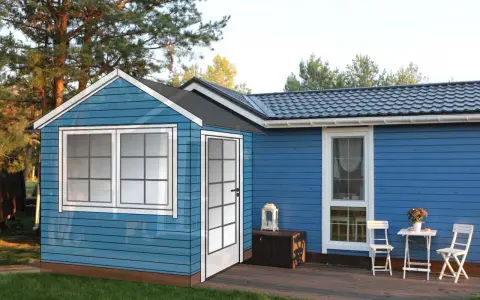 Blå trähus med fönster och dörr, redo att bygga farstu. En liten uteplats med bord och stolar.