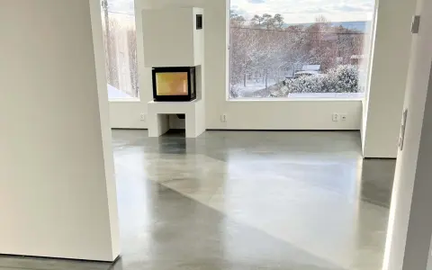 Ett modernt vardagsrum med en öppen spis och stora fönster som vetter mot en snötäckt trädgård. Golvet är ett vackert polerat betonggolv som reflekterar ljuset.