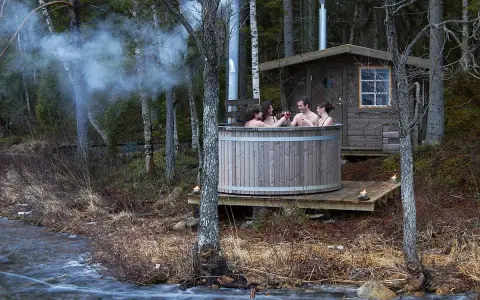 Fyra vänner njuter av en badtunna vid en sjö. Röken stiger från skorstenen, en mysig stund att bygga badtunna för.