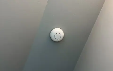 En vit, rund, uppkopplad brandvarnare sitter monterad i ett tak med två olika gråa nyanser.