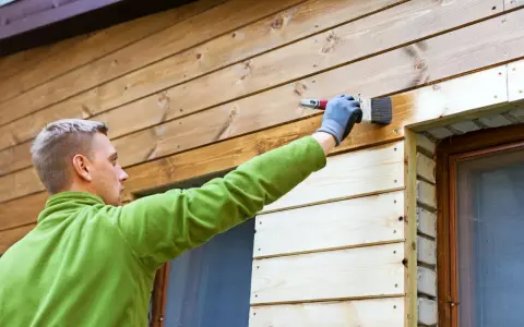 Man målar tryckimpregnerat trä med en pensel. Han har en grön tröja och grå handskar.