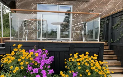 Vacker altan med glasräcke och trädetaljer. Blommor i gult och lila ramar in staketet. Perfekt altan staket inspiration för en modern uteplats.