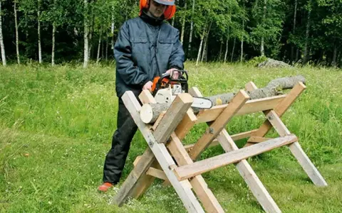 Man i skyddsutrustning använder motorsåg för att kapa ved på en sågbock. Ritning av en sådan konstruktion kan vara användbar.