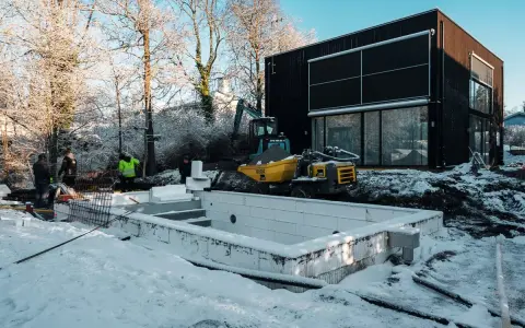 Snö täcker byggarbetsplatsen där en utbyggnad av husets långsida pågår. En grävmaskin och en dumper syns vid en nybyggd poolgrund.
