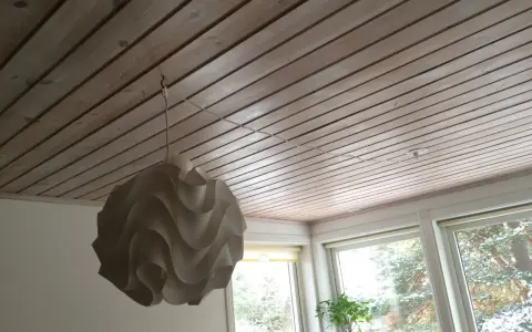 En vit taklampa hänger från en krok i ett träpanelat tak. En tunn sladd löper från lampan längs taket mot ett uttag.