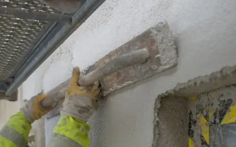 Händerna i gula handskar drar en träslev över en vägg, applicerar putsbruk b eller c för att skapa en jämn yta.