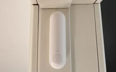 En vit, avlång sensor med en knapp sitter monterad på en ljusbeige dörrkarm. Den är en del av ett Aqara Thread-system för smart hem.
