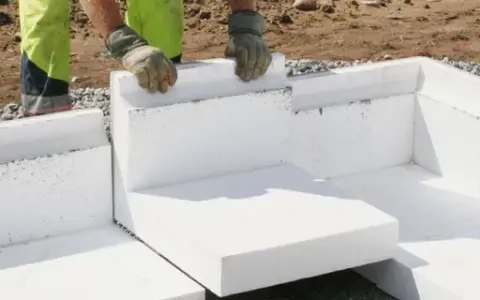 Arbetare monterar isolerblock för att gjuta platta garage. Grus och jord syns runt grunden.