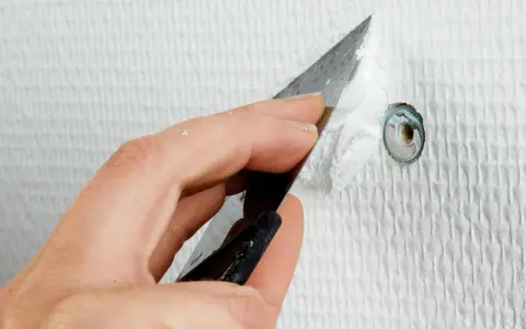 Hand spacklar ett litet hål i en vit, strukturerad vägg med en spackelspade.