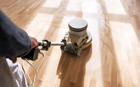 En person slipar ett trägolv med en maskin. Dammet virvlar upp och skapar ett mönster på det ljusa träet.