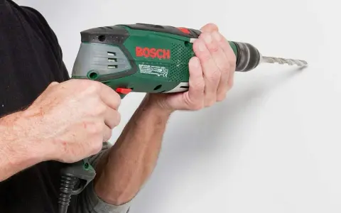 Händerna håller en grön Bosch-borrmaskin som borrar i en vit vägg.