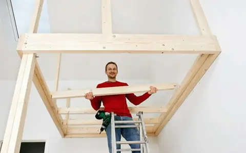 Man på stege bygger ett loft av trä. Han håller en träbit och ler.