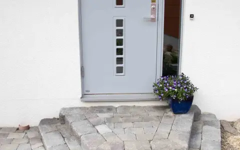 En vacker stentrappa leder upp till en modern ytterdörr. En blå kruka med blommor ger färg åt entrén.